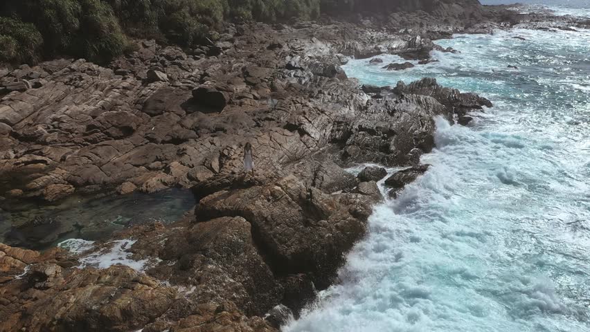 An epic drone shot showcases a bride standing on the rugged coastline of Sri Lanka, with waves crashing against the rocks. Ideal for promoting wedding destinations, adventure, and natural beauty