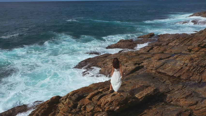 A breathtaking aerial view of a woman in a white dress standing on a rocky cliff, gazing at powerful ocean waves crashing against the rocks. Perfect for adventure and nature themes. 