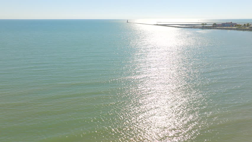 Drone aerial over Lake Michigan shimmering in bright sunlight toward the Sheboygan, Wisconsin lighthouse on the horizon on a beautiful clear day.