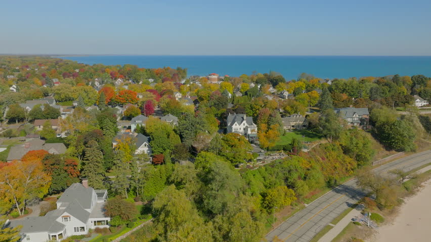Drone aerial panning over Sheboygan, Wisconsin neighborhood homes and autumn trees with Lake Michigan on the horizon under a clear, pretty blue sky.