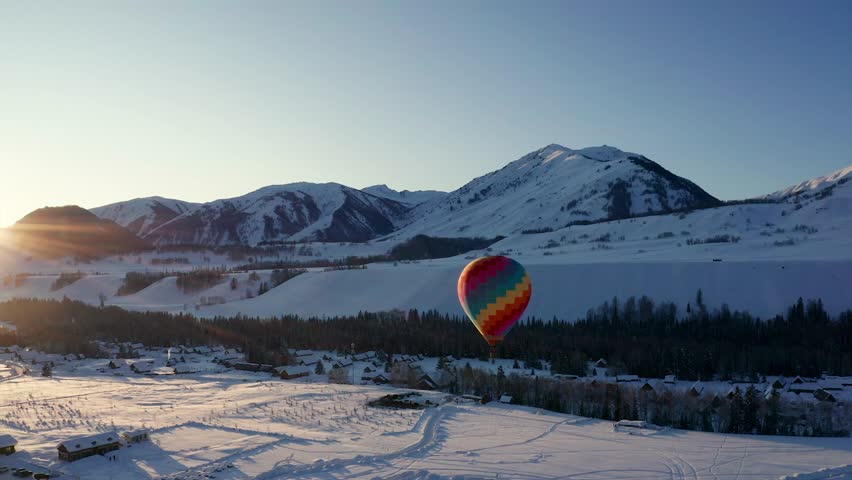 Colorful hot air balloon floating over snowy winter landscape with mountains and forest at golden hour sunset, creating a breathtaking aerial adventure scene.
