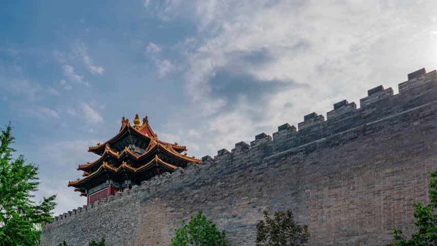 Ancient Chinese pagoda tower rising from stone fortification wall corner in Beijing, featuring traditional tiered architecture against cloudy blue sky with green foliage.