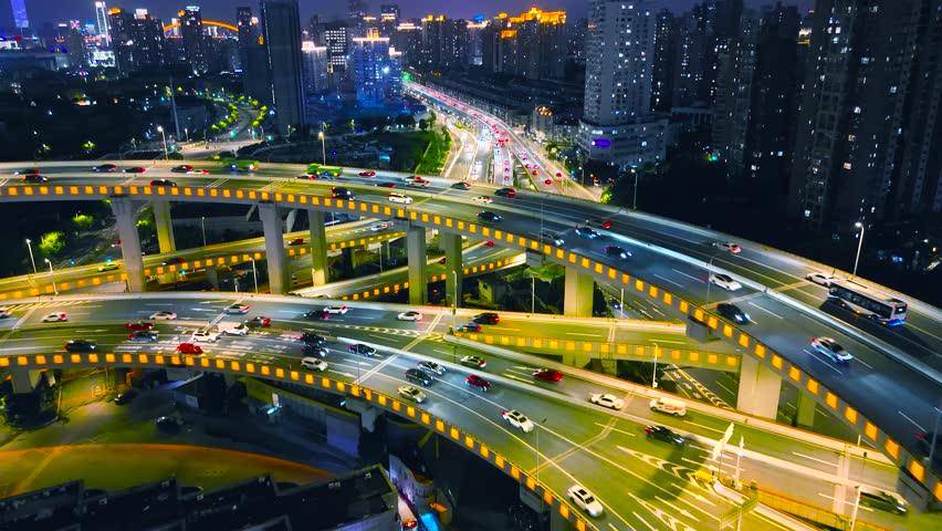 Aerial view of a complex highway interchange with illuminated traffic flowing through multiple road layers, surrounded by urban skyscrapers at night in Shanghai, China.