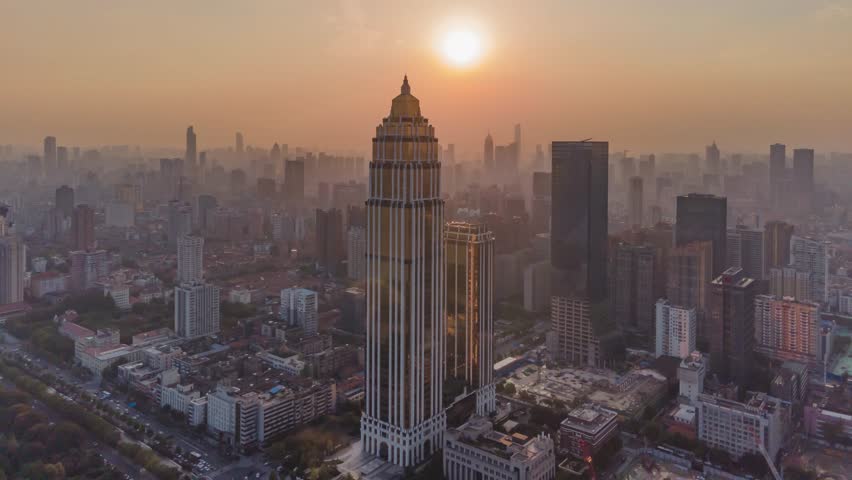 Aerial timelapse view of Wuhan cityscape at sunset with prominent landmark skyscraper and urban skyline bathed in golden light along the riverbank.