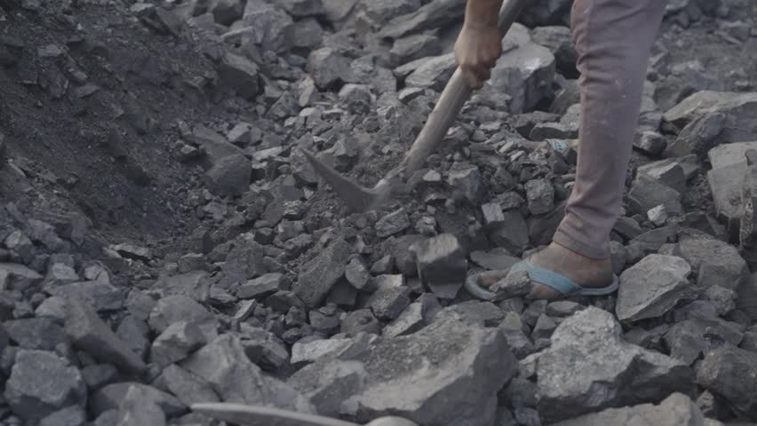 Closeup of a daily wage worker extracting coal at coal mine, wearing open toed sandal and pant, the individual is actively engaged in breaking or moving jagged pieces of coal using a hand tool
