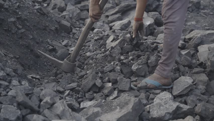 Young male worker extracting coal at coal mine, wearing open toed sandal and pant, the individual is actively engaged in breaking or moving jagged pieces of coal using a hand tool, Bokaro, Jharkhand