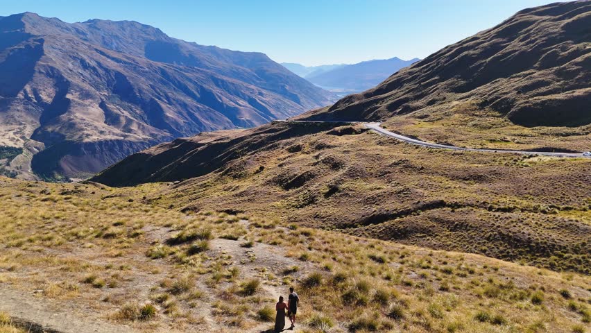 A couple walks through alpine scenery to enjoy views of Crown Range Road, the highest road in New Zealand. Mountains, winding roads, and valleys create a stunning natural scene.