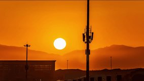 Cell phone tower silhouette against vibrant orange sunrise sky in desert landscape with mountains and buildings visible in background - Powered by Shutterstock - Get 15% off with code: PIKWIZARD15