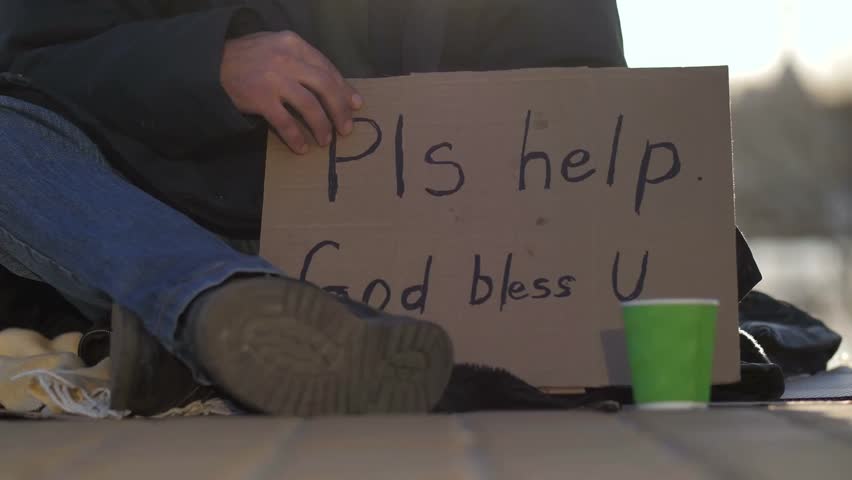 Close-up of a beggar’s hands and legs holding a cardboard sign asking for help, highlighting poverty, homelessness, social issues, and human struggle.