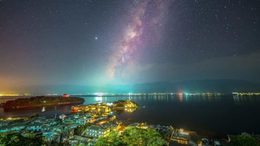 Stunning Milky Way galaxy stretching across starry night sky over illuminated coastal town and lake, creating magical astrophotography landscape with vibrant colors and reflections.