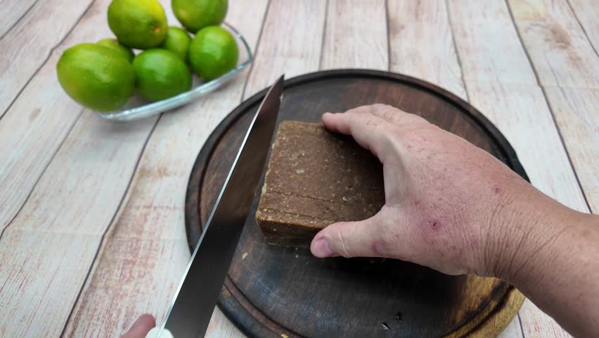 close-up shot hands using a knife to slice a block of panela on a rustic wooden board, ready for a sweet recipe sugar cane limeade