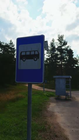 Vertical, low-angle shot focusing on a blue and white bus stop sign post with a bus silhouette, contrasting against a cloudy blue sky. A dirt road and a rustic metal shelter are visible in the pine