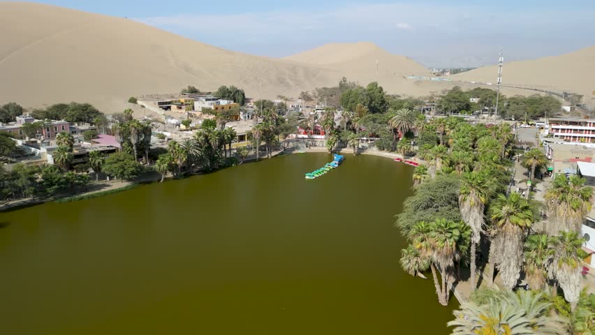 Aerial parallax shot of the Huacachina desert oasis with lake and buildings in Ica, Peru, surrounded by palm trees and large sand dunes.