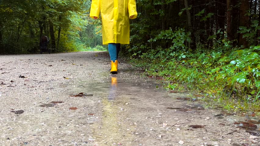 Woman with yellow rubber boots and raincoat walking through puddles in the park on a rainy autumn day.