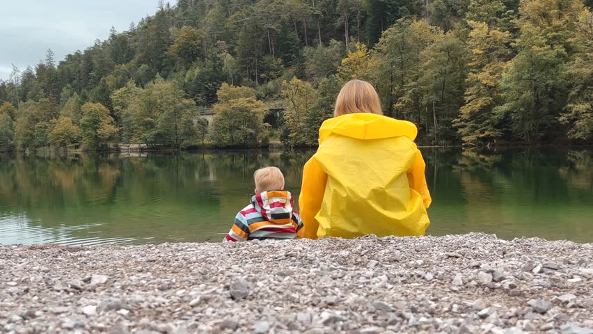mother and son in raincoats sit on the shore of a park lake and throw pebbles into the water. Family time in nature, precious moments in life.