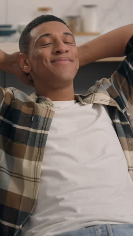Relaxed carefree smiling African American man guy male at home relaxing in living room leaning back on couch comfortable sofa enjoying new home own tenant weekend indoors break pause thinking dream