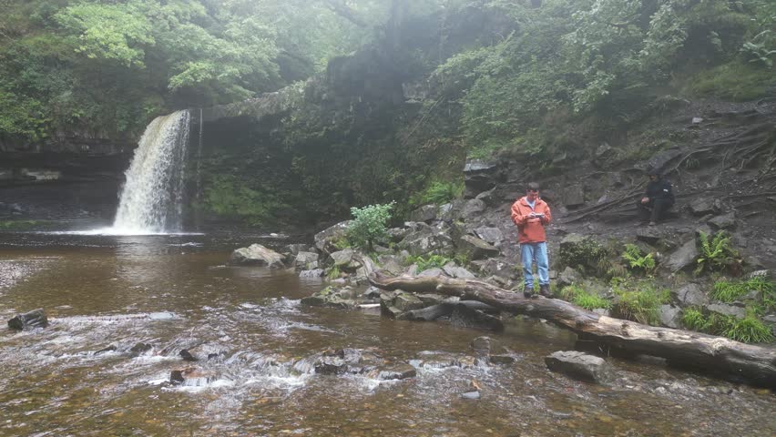 Drone footage of a young man standing by Angel Falls in Brecon Beacons National Park on a cold, wet day, gazing away from the cascading waterfall, surrounded by rugged rocks and lush green forest.