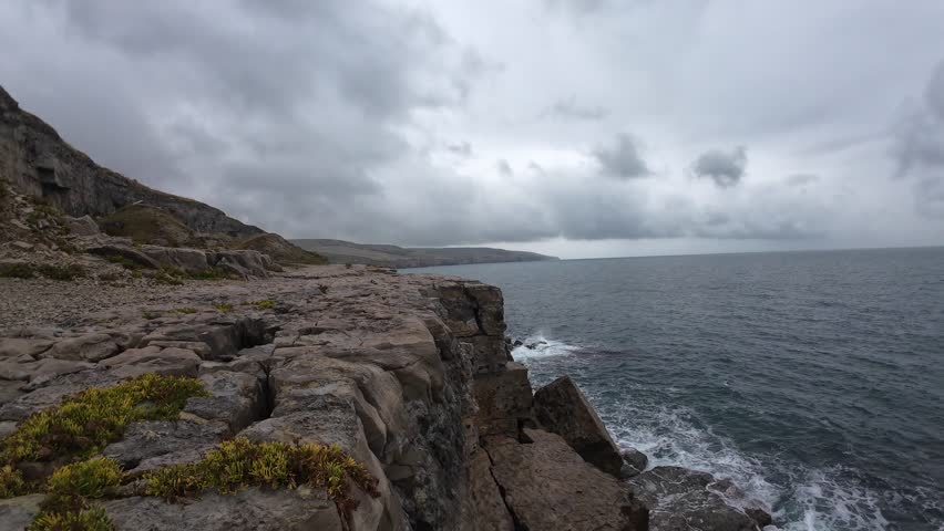 Drone footage captures dramatic waves crashing against rugged ledge rock formations in Dorset's Isle of Purbeck. Moody skies and powerful sea evoke nature's raw power.