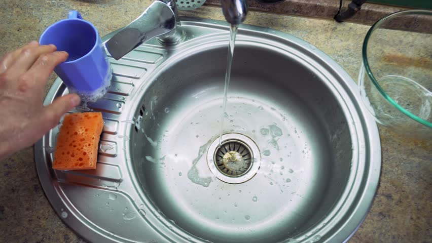 Man rinsing blue mug under running water to remove soap suds in kitchen sink