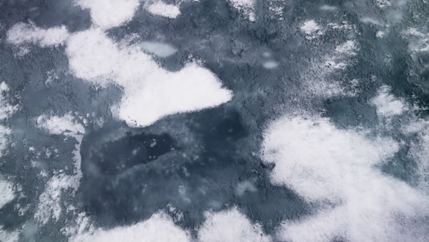 Aerial view of a frozen lake transitioning from scattered snow-covered ice floes to fractured, jagged plates, revealing deep blue cracks and textures as winter surface thaws.