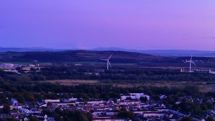 Aerial view of a peaceful town at dusk, with glowing sunset skies, wind turbine, distant hills, and warm twilight tones blanketing rooftops and countryside in calming purple hues