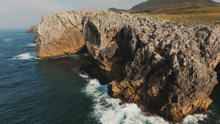 Dramatic rocky cliffs rising above the Atlantic Ocean, with waves crashing at their base on a rugged coastline.