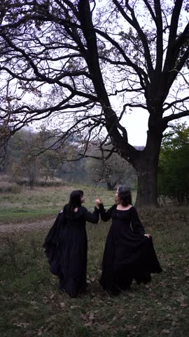 Two women in long black dresses perform a historical reenactment of the Edwardian era dancing lovers on a hill near a majestic oak tree for Halloween.