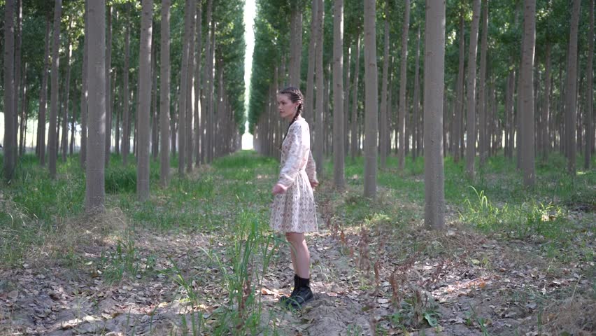 A young woman stands gracefully in a serene forest, wearing a beautiful white dress that contrasts elegantly with the lush backdrop of tall trees and vibrant greenery surrounding her