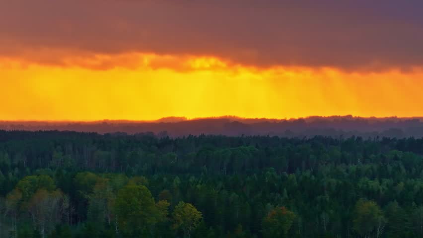 Vivid orange sunset casts golden light on distant rain over a dense forest canopy in Europe