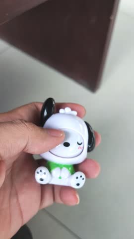 Hand holding a cute white plastic figure (likely Sanrio Pochacco or similar) with a blinking or winking expression. Captures the playful mechanism and toy detail in a bright interior setting.