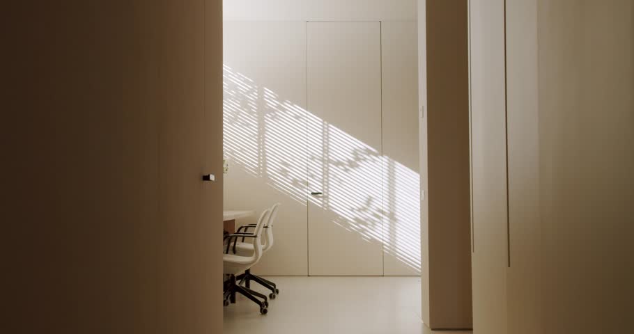 Sunlight streams through window blinds, casting shadows in a modern, minimalist office space. White chairs sit beside a simple table, Natural light illuminates the room, work