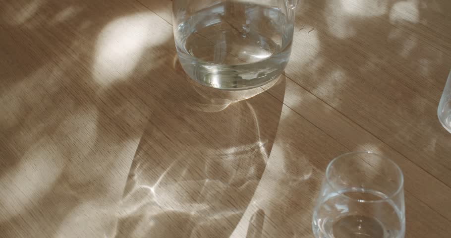 Sunlight streams through a window, casting dancing shadows on a wooden table where a glass pitcher of water sits beside two clear glasses. One glass is empty, the other half full,