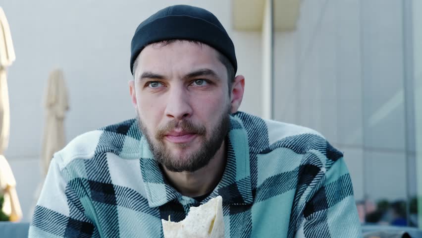 Young man in a checkered jacket and black cap eating a burrito or shawarma outdoors. Horizontal 4k footage