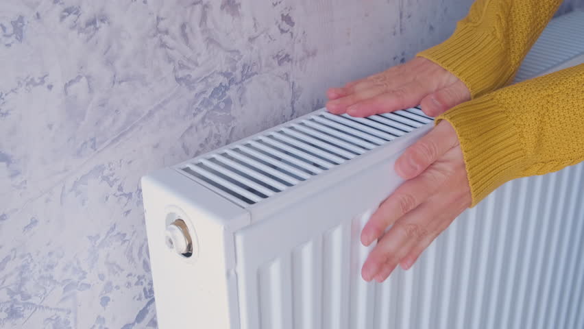 Closeup of man warming his hands in yellow sweater on the heater at home during cold winter days. Male getting warm up his arms over radiator. Concept of heating season or cold weather