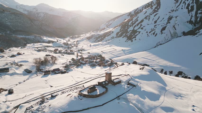 Aerial pan view Ancient Lamaria Monastery in snowy highland valley with Ushguli settlement village houses in cold winter land surrounded by Caucasus mountains. UNESCO - listed Upper Svaneti region
