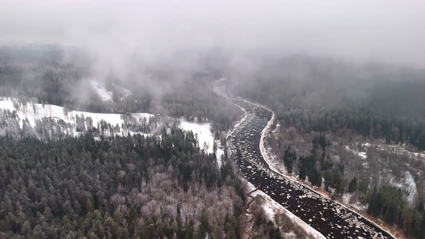 Aerial pullback through fog above snowy forest and ice covered winding winter river