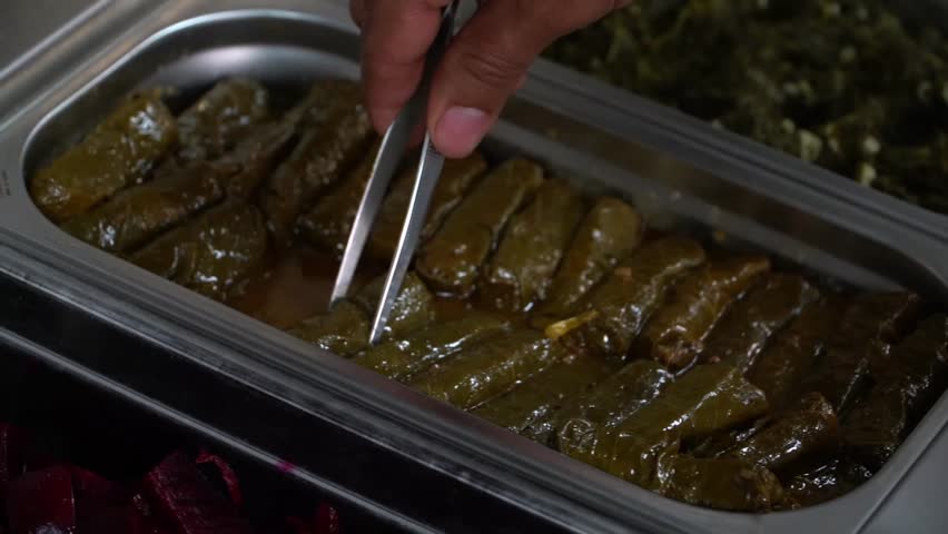 A close-up image of stuffed grape leaves in a stainless steel serving tray with some green leaves and beets in the background.