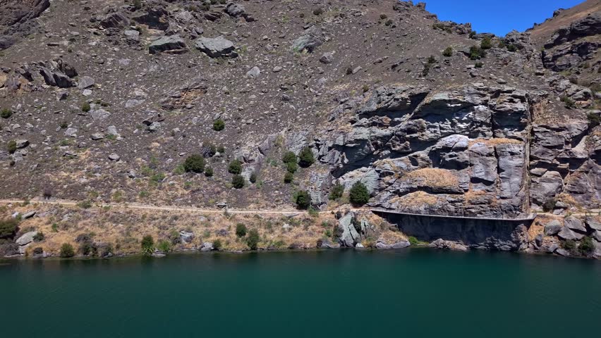 Aerial sideways drone view following the Dunstan Trail on a sunny day, showcasing rugged rocky hills and the Clutha River’s vivid blue waters in Central Otago, New Zealand.