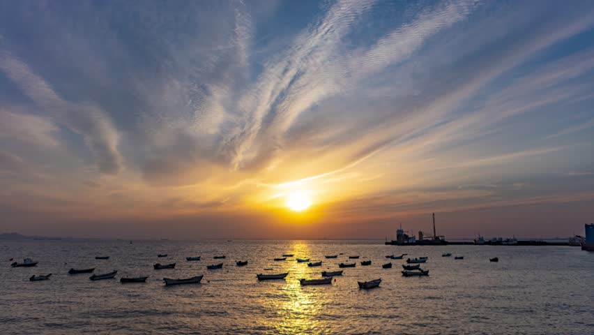 Beautiful sunset time lapse over fishing harbor with boats anchored in calm waters under dramatic streaming clouds during golden evening hour