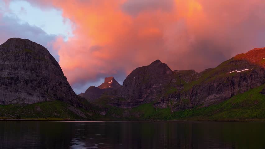 Dramatic mountain range reflected in calm lake waters during colorful sunset evening, creating stunning time-lapse worthy landscape with vibrant orange and pink sky colors.