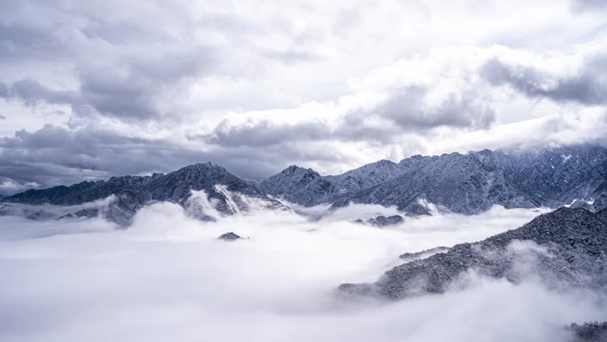 Dramatic mountain peaks emerging from misty clouds with snow-capped summits under stormy sky, creating ethereal landscape scene perfect for nature and travel content.