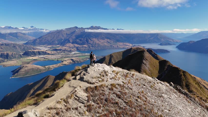 A 4K drone pans around a couple at the iconic Roy’s Peak viewpoint in Wanaka, New Zealand. Scenic mountains, lakes, and alpine trails make up the stunning views from the top of the vantage point.