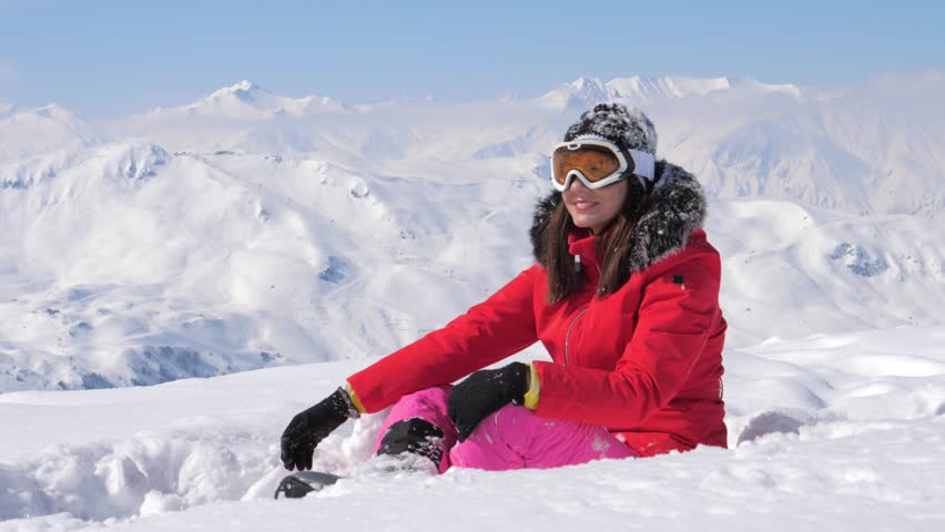 Young woman sitting on top of snow mountain, smiling at camera and gesturing thumbs up and ok sign while on winter vacation at beautiful ski resort in the alps