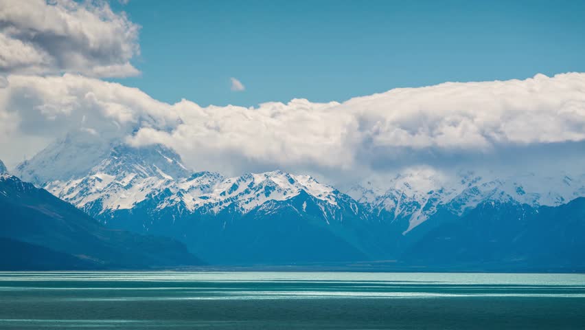 Dramatic snowy mountain peaks with mist and clouds over tranquil alpine lake, creating stunning natural landscape with blue sky and serene water reflections.