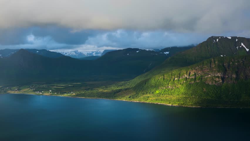 Dramatic timelapse view of pristine mountain lake surrounded by snow-capped peaks and lush green forests under stormy clouds with rainbow light breaking through