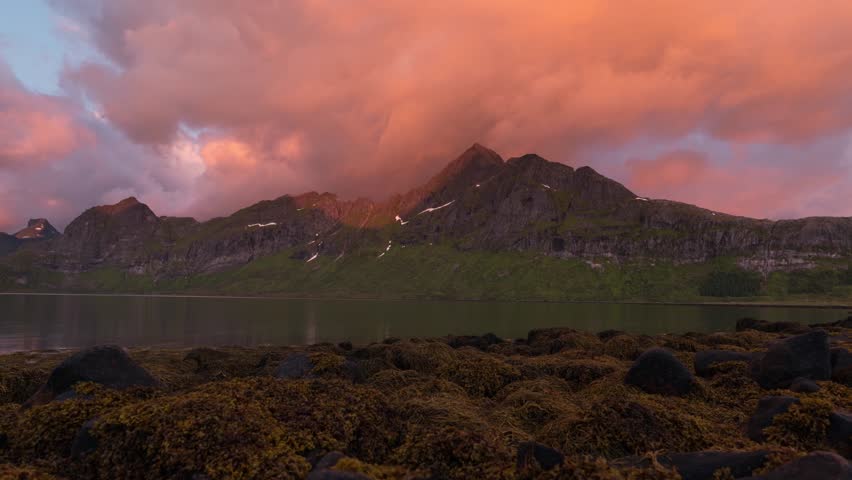 Dramatic mountain range reflected in calm lake waters during golden sunset with colorful evening sky and rocky shoreline in foreground
