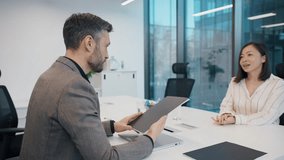 Man in blazer reading documents during meeting with Asian woman in striped blouse at white desk. Professional office environment with glass walls and modern furniture. New employee interview. CV. - Powered by Shutterstock - Get 15% off with code: PIKWIZARD15