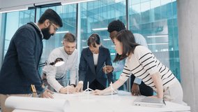 Diverse group of young professionals leaning over desk discussing project layout in modern office. Multinational team analyzing wind turbine model and blueprints near window. Indoors. Teamwork. - Powered by Shutterstock - Get 15% off with code: PIKWIZARD15