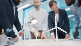 Close up wind turbine model standing on table with blurred diverse business team brainstorming in background. Multiracial group working on renewable energy project in modern office. Indoors. - Powered by Shutterstock - Get 15% off with code: PIKWIZARD15