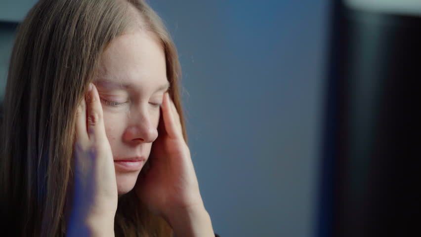 Tired middle-aged Caucasian woman studing in front of desktop computer fresh face, massaging her temples. Close-up, minimalist background, dark room, dimly lit.